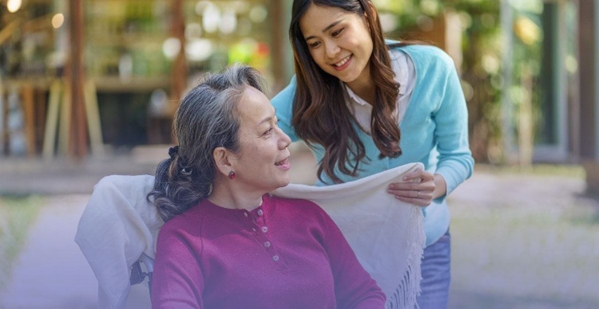 A smiling caregiver gently placing a blanket over an older adult seated outdoors, representing comprehensive care and protection for loved ones with incontinence.
