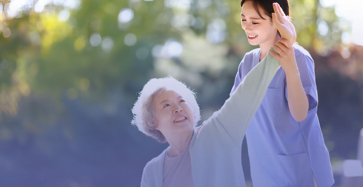 A smiling young female caregiver assisting an older woman with gentle arm movements outdoors.