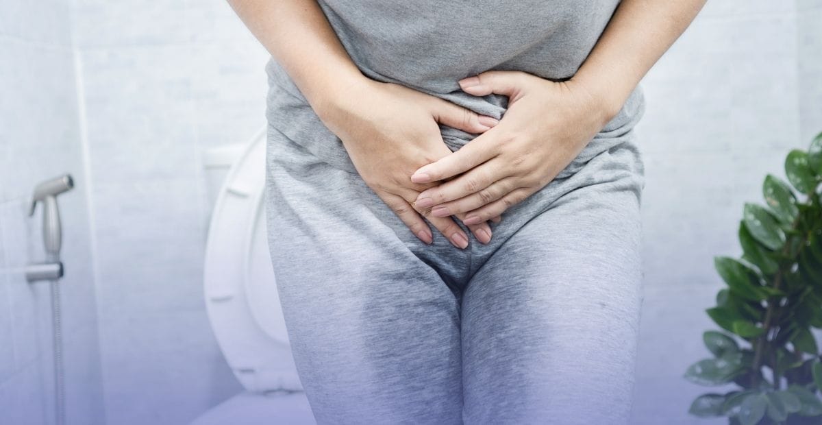A person holding their lower abdomen near a toilet, representing symptoms and causes of urinary incontinence.